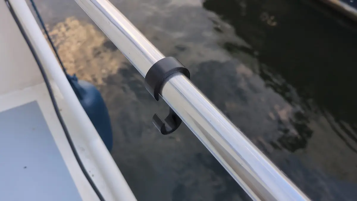 Nahaufnahme eines schwarzen Kunststoff-Relinghakens, der an einer silbernen Bootsreling befestigt ist. Der Haken dient als Halterung für Landstromkabel oder Schläuche und ist bereit zur Anwendung, während im Hintergrund das Wasser und ein blauer Fender zu sehen sind.