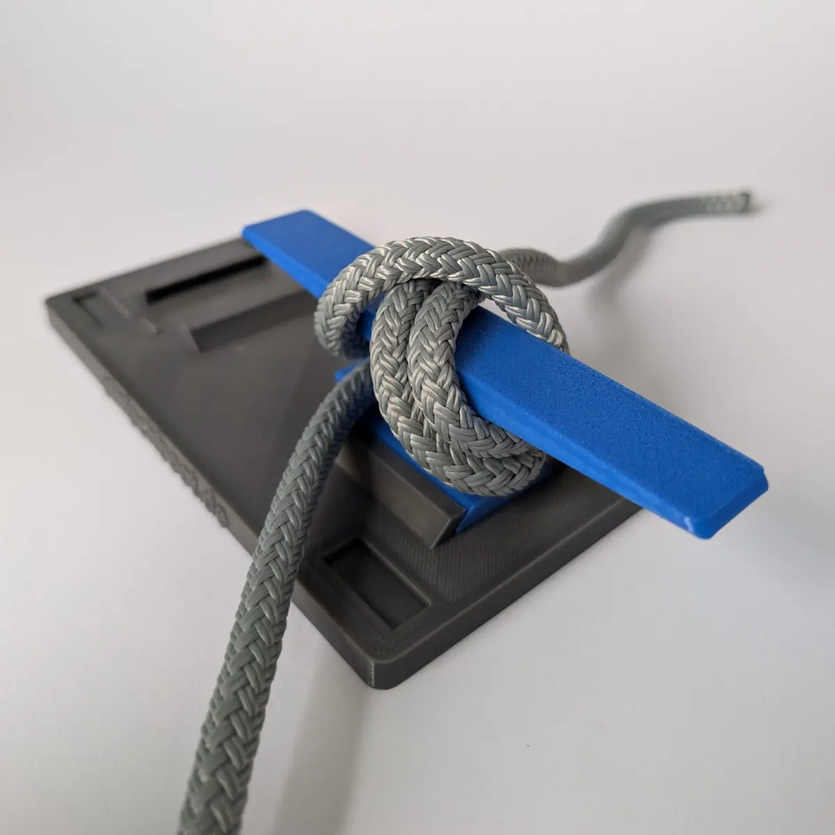Eine graue Basisplatte mit einer blauen Klampe, um die ein graues Seil (Tauwerk) mit einem Seemannsknoten befestigt ist. Dies zeigt die vielseitige Übungsmöglichkeit von Knoten auf dem Knotenbrett.