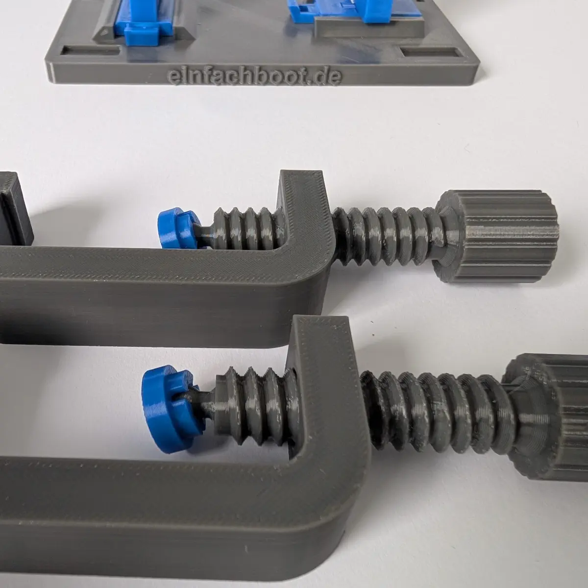 Nahaufnahme von zwei grauen Schraubzwingen mit blauen Druckstücken aus Kunststoff. Die Zwingen zeigen ein robustes Gewinde zur flexiblen Befestigung des Knotenbretts an einer Tischplatte