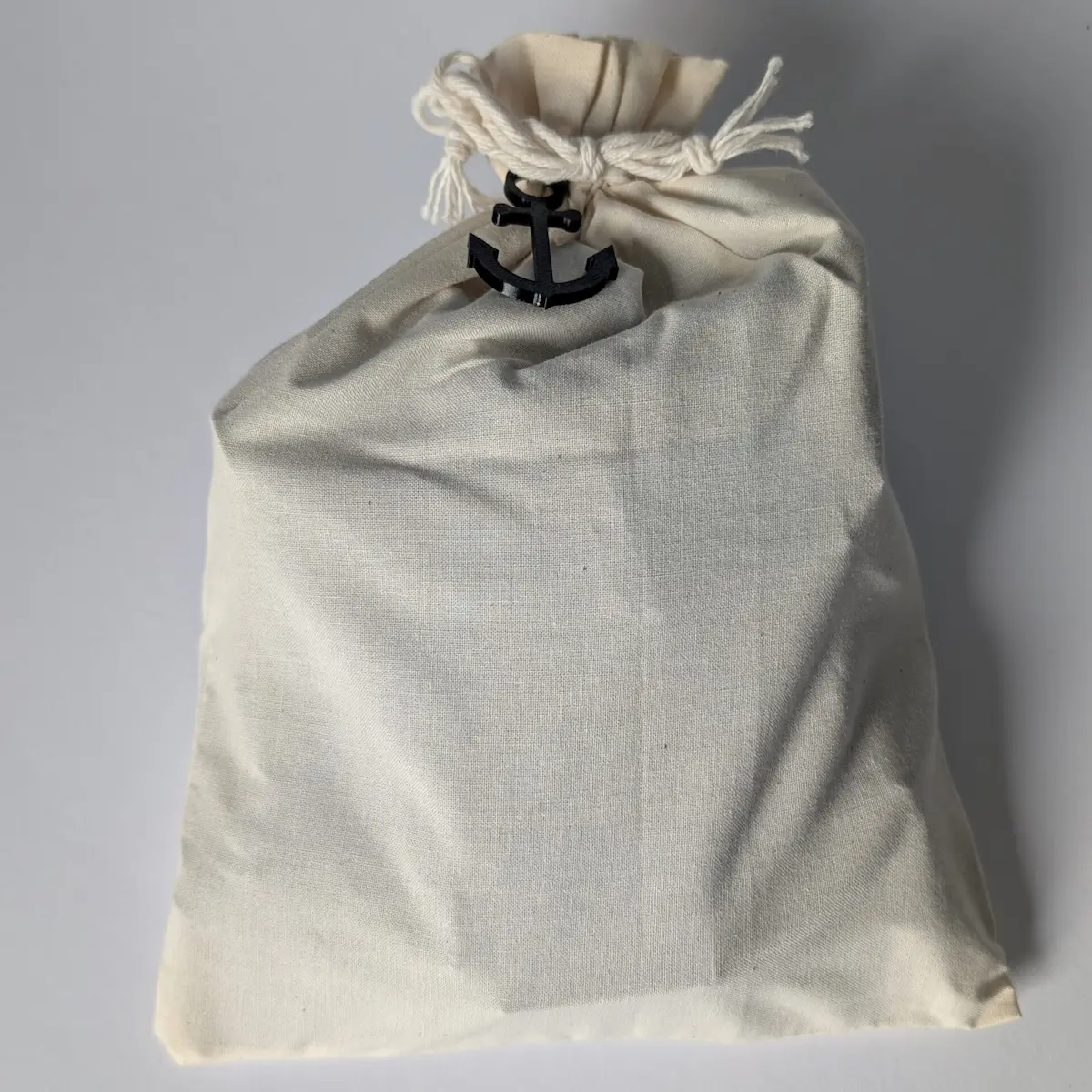 Hochwertige Geschenkverpackung für das Knotenbrett: Ein cremefarbener Baumwollbeutel mit Kordelzug, verziert mit einem kleinen schwarzen Deko-Anker.
