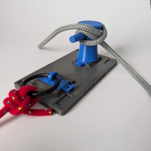 Graue Basisplatte mit zwei blauen Modulen: Ein kleiner Poller, um den ein graues Seil belegt ist, und ein schwarzer Ring an einer blauen Halterung mit einem roten geknoteten Seil.